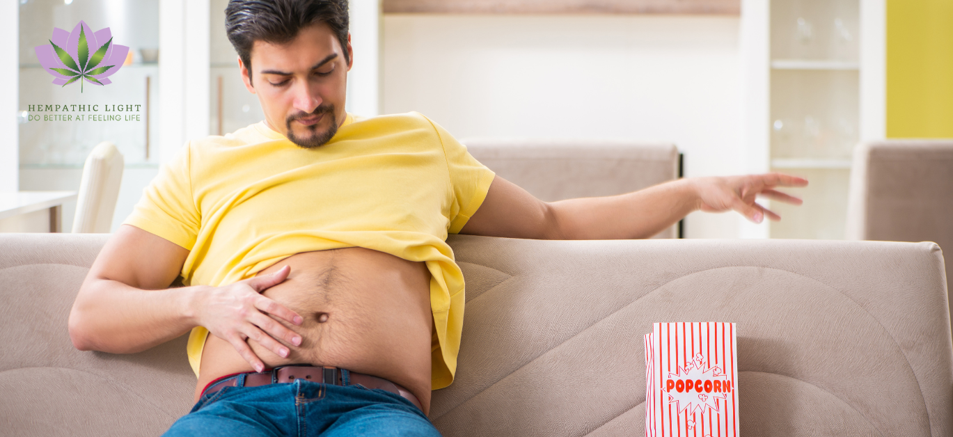 Homem sentado no sofá, com a t-shirt arrgaçada a mostrar o estômado e a mão direta a afagar a barriga. Sugere incómodo nessa zona.